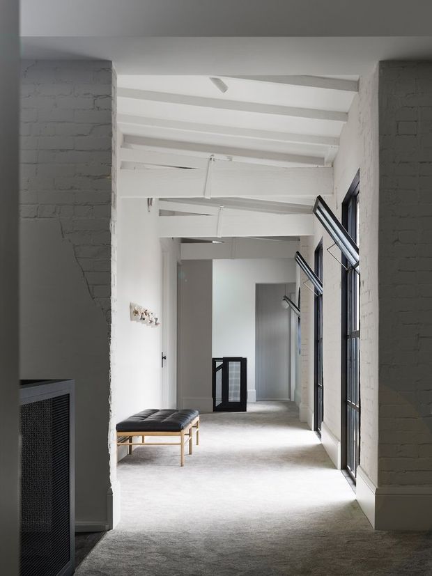 A long, narrow interior corridor or gallery with white-painted exposed brick walls, exposed timber beams and rafters, and tall black-framed windows along one side. The space has pale carpeted flooring, a simple upholstered bench, minimal wall lighting, and strong natural light, creating an airy, restrained industrial character.
