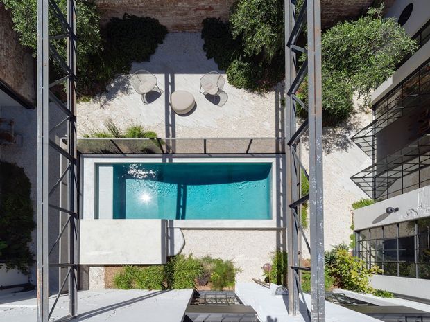 A high-angle view into a contemporary courtyard framed by dark metal structural elements and surrounding buildings. The space includes a rectangular turquoise swimming pool, pale paved surfaces, sculptural outdoor lounge chairs, clipped greenery, and exposed brick walls, creating a geometric indoor-outdoor composition.