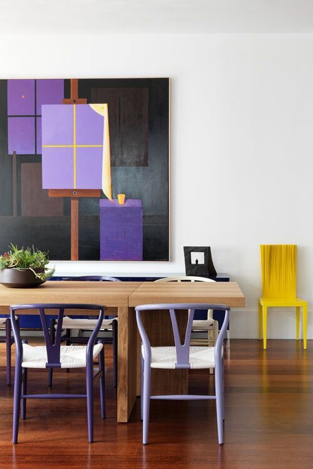 A refined dining area with a long timber dining table and purple upholstered dining chairs on polished timber flooring. A large abstract artwork in purple, yellow, brown, and black hangs above the table, complemented by a bright yellow sculptural chair and a small plant. The room has white walls, contemporary furniture, and a strong, colourful modernist palette.
