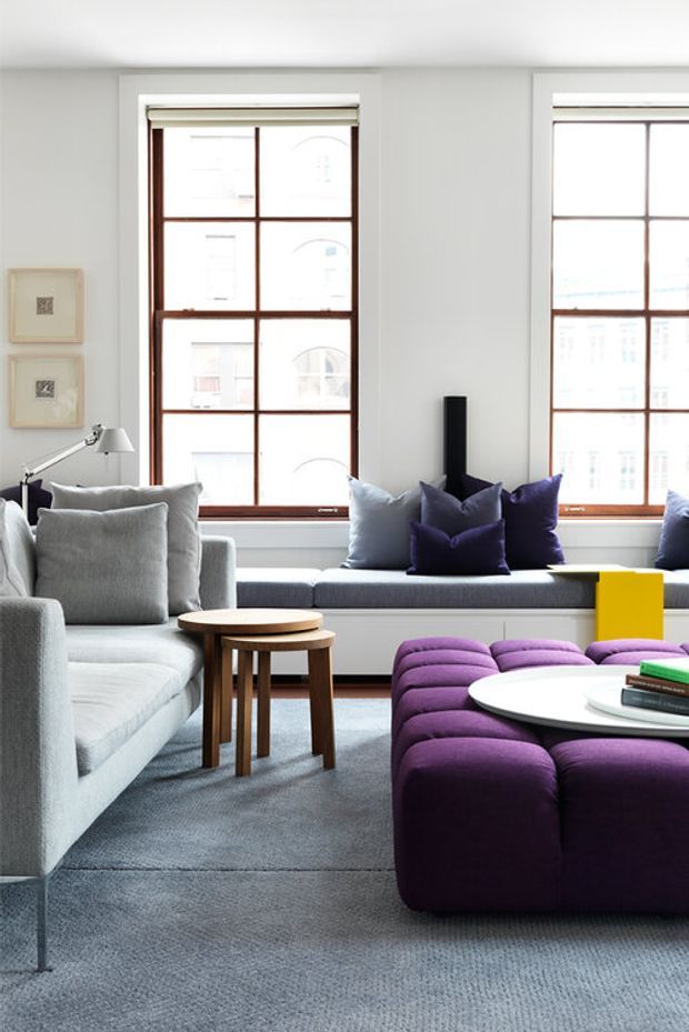 Bright, contemporary living room with large timber-framed grid windows, a built-in window seat, pale grey sectional sofa, purple upholstered ottoman seating, nested timber coffee tables, and a small yellow side table. The room has a soft grey carpet and a clean, refined palette with bold purple and yellow accents.