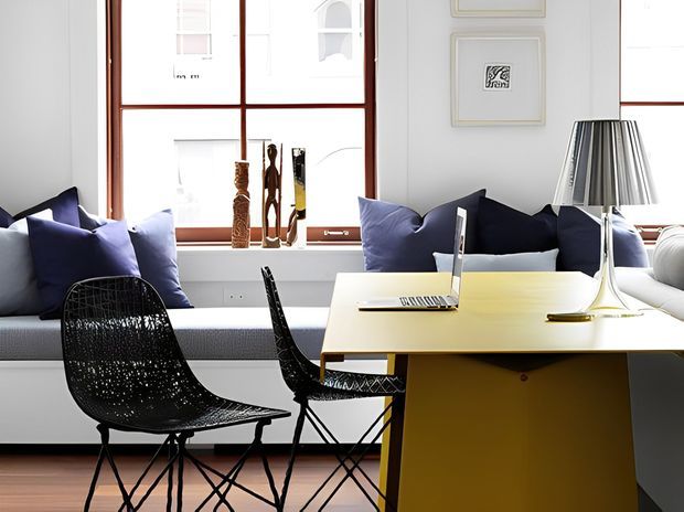 Bright, stylish open-plan living and dining area with a built-in window seat layered with navy and pale cushions, a sculptural black woven lounge chair, a compact black dining chair, and a mustard-yellow rectangular dining table. Large timber-framed windows, framed artwork, decorative objects, and a contemporary table lamp create a refined mid-century-inspired interior.