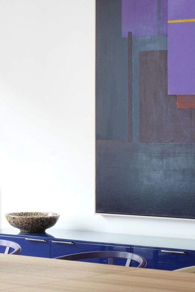 A close-up view of a contemporary dining area, featuring a large abstract artwork with deep blue, purple, teal, and muted red geometric forms on a white wall. Below it is glossy deep-blue built-in cabinetry with slim metal hardware, a stone bowl, and the edge of a light timber dining table with curved dark chairs in the foreground.