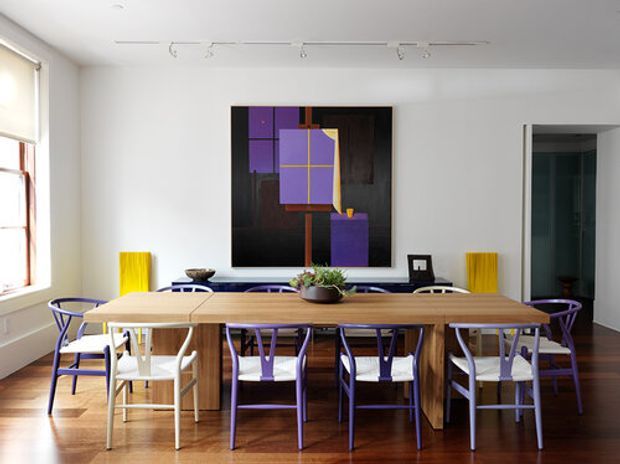Bright contemporary dining room with a long natural timber dining table surrounded by mismatched chairs, including vivid purple and white seating. White walls, timber flooring, track lighting, a large abstract artwork, and tall windows create a clean, gallery-like setting. A doorway at right leads to another room.