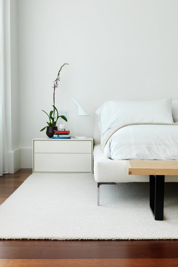 A bright, minimalist bedroom vignette featuring a low upholstered bed with crisp white bedding, a pale upholstered headboard, and a simple timber bedside bench. A white two-drawer nightstand holds a potted orchid and stacked books, with a small white wall-mounted reading light above. A large white rug sits over dark timber flooring, while soft white walls and restrained furnishings create a clean, serene composition.