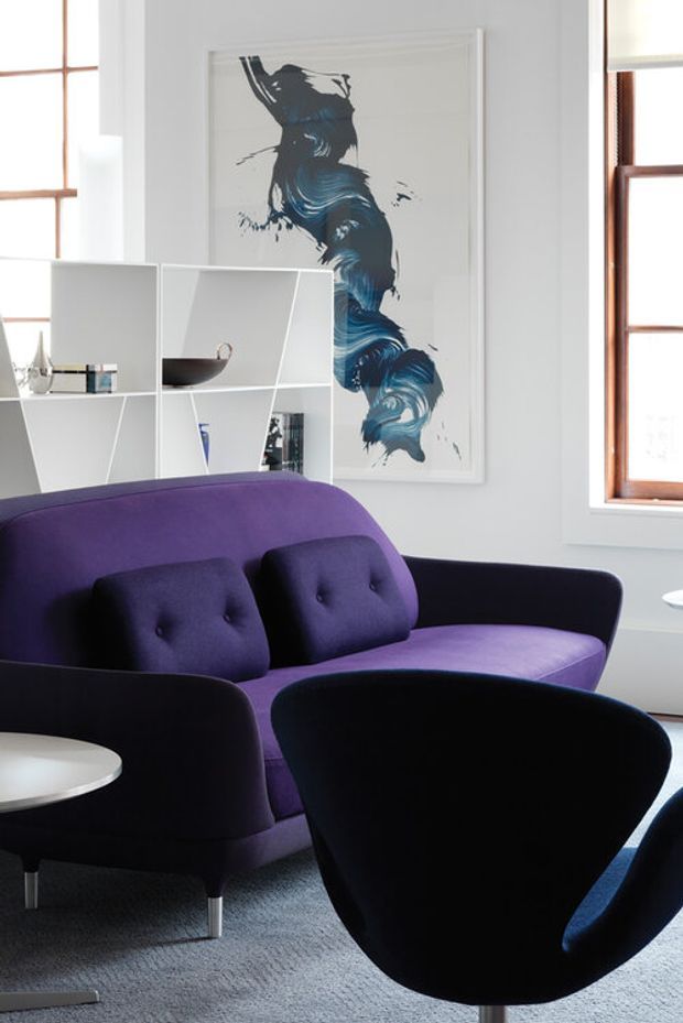 A bright, stylish living room featuring a deep purple upholstered sofa with button-tufted cushions, matching dark lounge chairs, and a small round side table. A white geometric open shelving unit displays books and decorative objects, while a large abstract blue artwork and timber-framed windows add visual interest. The room has a soft grey carpet and a clean, contemporary arrangement.