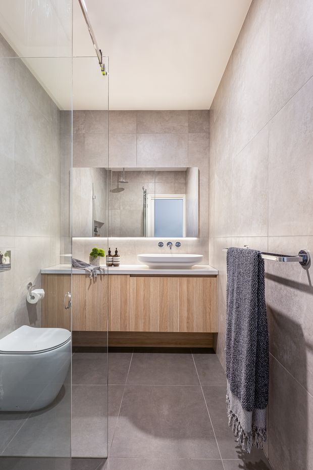A compact contemporary bathroom with full-height grey-beige porcelain wall and floor tiles, a frameless glass shower enclosure, wall-mounted timber-look vanity cabinetry, white engineered-stone benchtop, vessel basin, mirrored splashback, wall-mounted tapware, and a modern white toilet. A textured dark grey towel provides contrast.