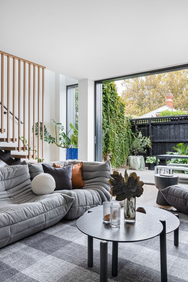 Bright contemporary living room with a large grey modular sectional sofa, round black coffee table, layered grey rug, and indoor plants. A timber-slatted stair screen and staircase are visible on the left, while full-height black-framed glazing opens to a leafy paved courtyard with a dark fence and outdoor furniture.
