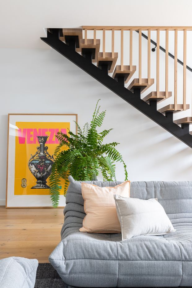 Bright contemporary living room featuring a large pale grey sectional sofa with neutral cushions, a timber floor, a black open-riser staircase with light timber balustrade, a large leafy fern, and colourful framed artwork leaning against the wall.