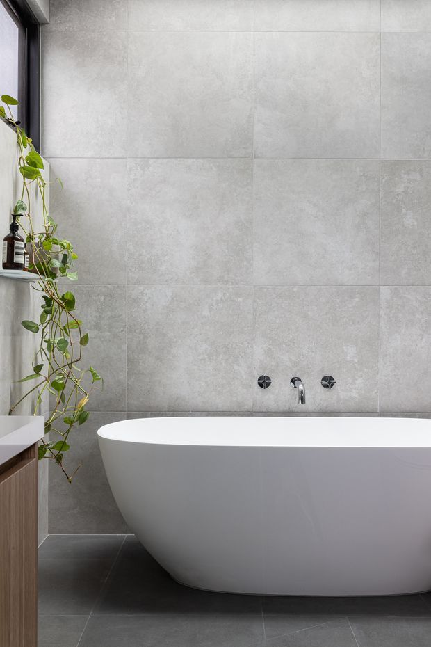 A contemporary bathroom featuring a freestanding white bathtub against large-format grey concrete-look wall tiles. Dark grey floor tiles, matte black window framing and tapware, and a trailing indoor plant create a restrained, modern palette. A partial timber vanity is visible at the left edge.