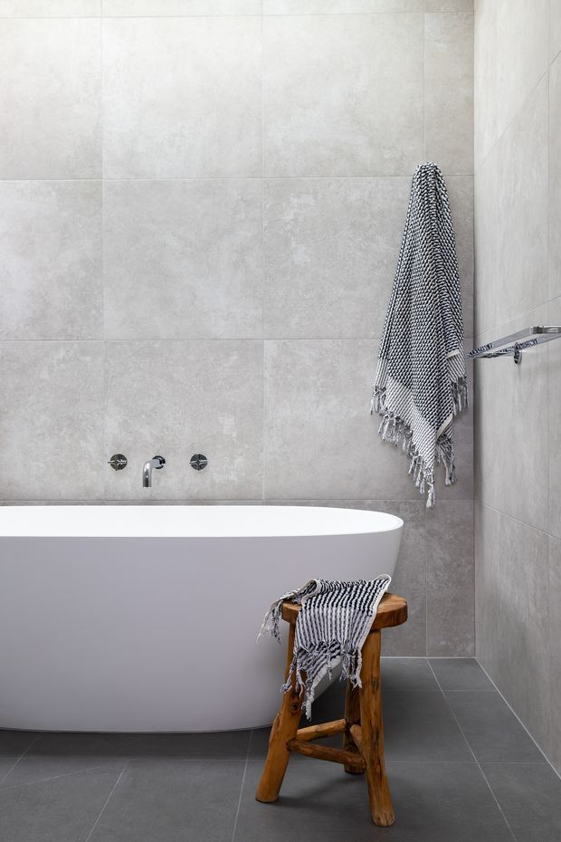 A minimalist contemporary bathroom featuring a freestanding white bathtub, large-format light grey wall tiles, dark grey floor tiles, and chrome tapware. A black-and-white patterned towel hangs from a wall-mounted rail, with another draped over a rustic wooden stool beside the bath.