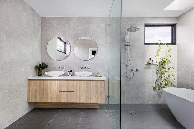 Contemporary bathroom with a floating light-wood double vanity, twin round mirrors, large-format grey wall and floor tiles, and a frameless glass shower enclosure. A freestanding white bathtub sits beside the shower, with a small high window and recessed shelf adding natural light and greenery.