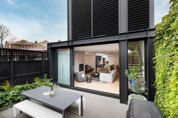 Contemporary black-clad rear extension opening onto a paved outdoor patio, with large glazed sliding doors revealing a bright living room inside. The facade features prominent dark horizontal louvres, black framing, vertical planting, a timber privacy screen, and outdoor dining furniture.