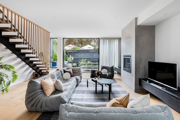 Bright contemporary living room with a large curved sectional sofa, additional lounge seating, a black coffee table, television and built-in fireplace set into a textured grey feature wall. The room has pale timber flooring, extensive glazing with sheer curtains, and an open staircase with timber balustrade and dark timber or metal treads.