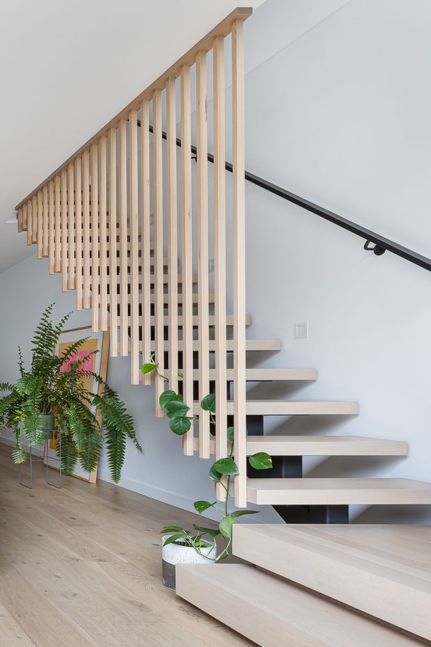 Bright contemporary interior view of a floating staircase with pale timber treads and a distinctive vertical timber slat balustrade. A black handrail runs along the white wall, while indoor ferns and trailing plants soften the stair hall. Pale timber flooring and white walls create a clean, airy look.