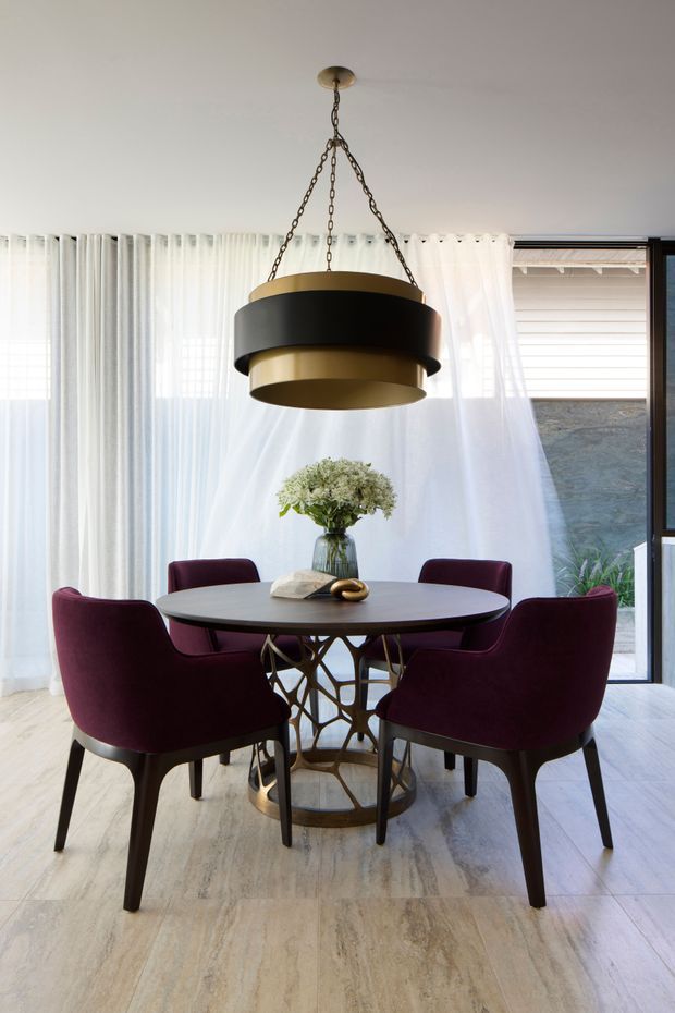 A refined dining area with a round dark dining table surrounded by four deep burgundy upholstered chairs. A sculptural black-and-brass pendant light hangs above, with sheer white curtains covering large windows and doors. The room has pale stone-look flooring and a bright, elegant atmosphere.