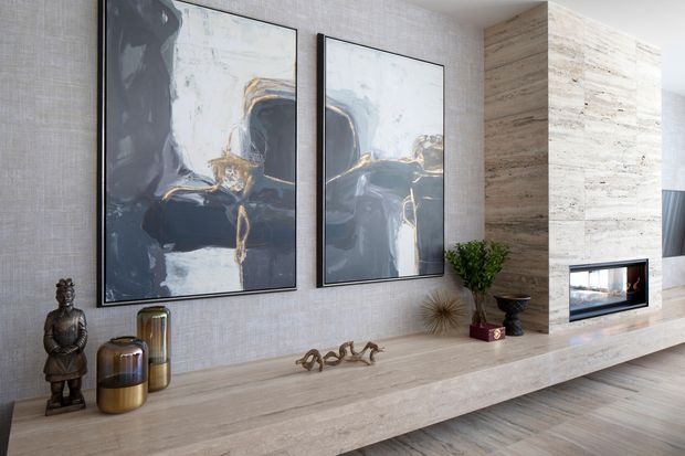 Contemporary living room feature wall with a long pale stone or travertine-like built-in console and fireplace surround. The textured neutral wall is decorated with two large abstract artworks, metallic vessels, a sculptural ornament, greenery, and dark decorative objects. A linear fireplace is integrated into the stone-clad volume, with timber flooring and soft natural tones creating a refined, warm interior.