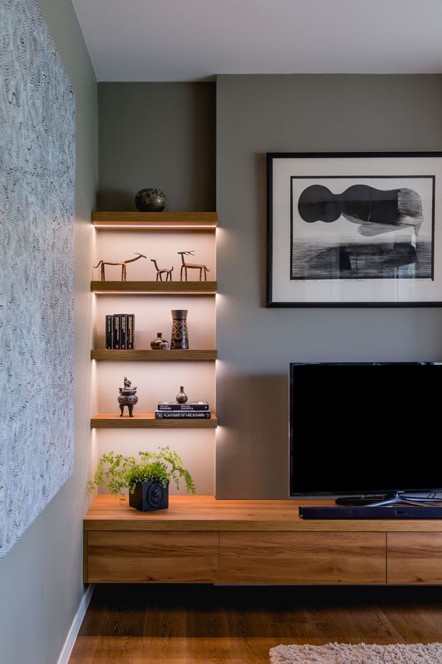 A refined living room media wall featuring a wall-mounted television, low walnut-toned built-in cabinetry, and illuminated open display shelving with books and decorative objects. The room has dark timber flooring, a warm grey painted wall, a large framed monochrome artwork, and a textured patterned feature wall at left.