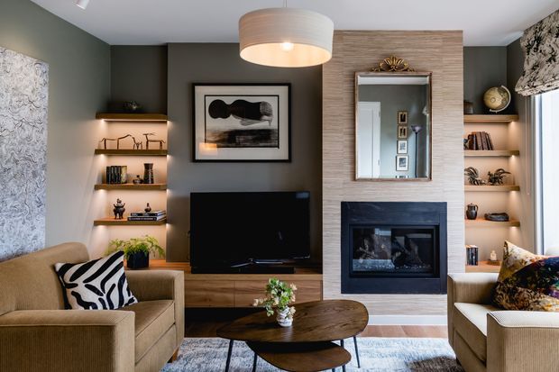 A warm, contemporary living room centered on a dark built-in fireplace and timber feature wall. The room includes a television and low timber media cabinetry, illuminated open display shelving, two upholstered sofas, a sculptural coffee table, patterned cushions, framed artwork, and a large mirror above the fireplace. Dark green-grey walls contrast with warm wood, stone-like fireplace cladding, and soft neutral furnishings.
