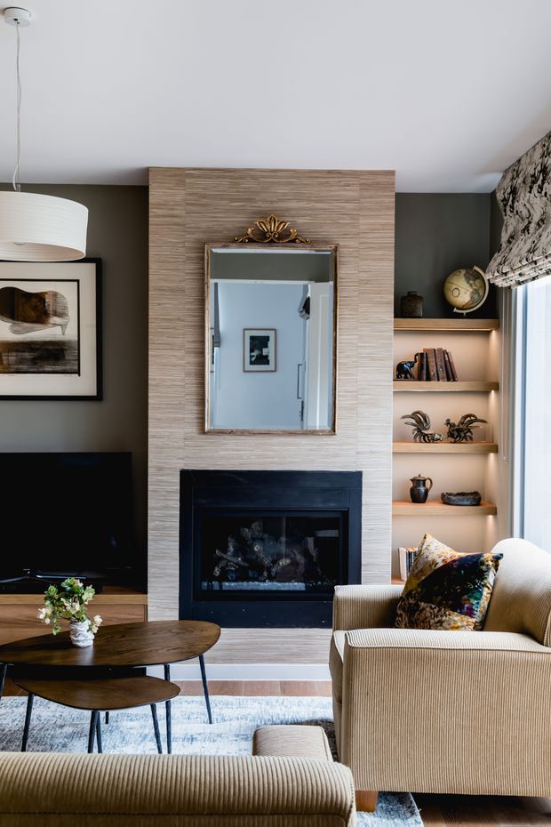 A refined living room centered on a dark built-in fireplace with a pale textured feature wall and decorative mirror above. Built-in open shelving displays books, ceramics, plants, and a globe beside the fireplace. The room includes a television, round wood coffee tables, ribbed upholstered armchairs and ottoman, patterned cushions, artwork, and a large pale rug. A window with patterned Roman blind provides natural light.