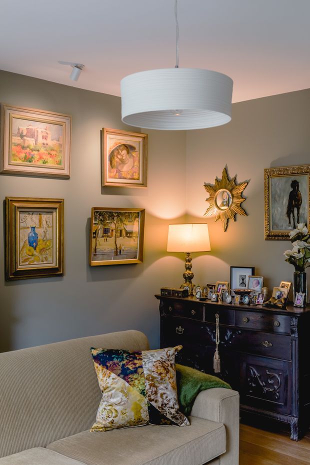 A warm, richly decorated living room corner featuring a taupe-painted wall covered with framed artwork, an ornate sunburst mirror, and a dark carved timber dresser displaying framed photographs and decorative objects. A ribbed white drum pendant light hangs above, with a table lamp providing ambient illumination. The foreground has a light upholstered sofa with colourful patterned cushions, and timber flooring is visible.