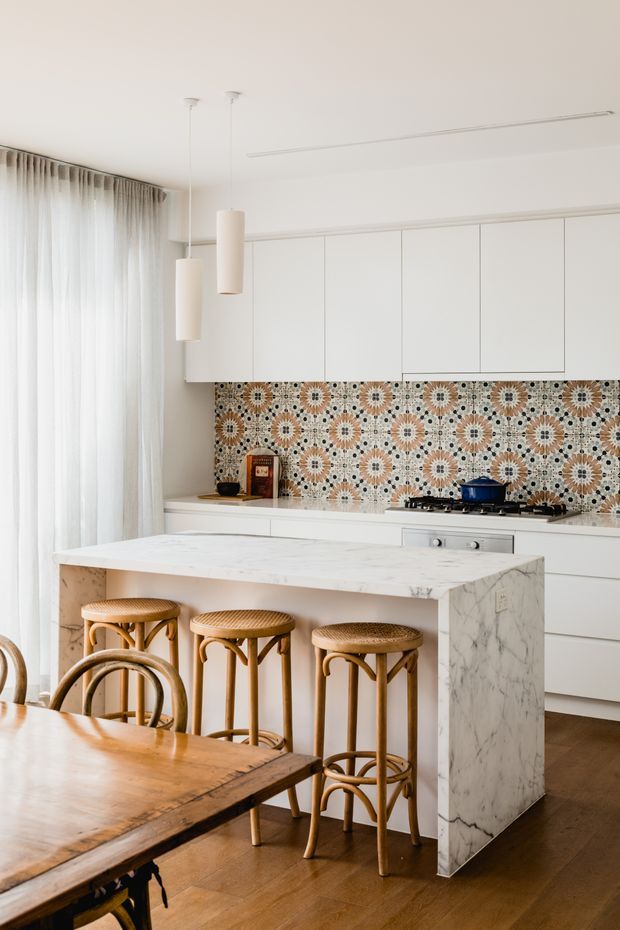 Bright, refined kitchen-dining interior with white flat-panel cabinetry, a large white marble island, patterned geometric tile splashback, timber flooring, woven rattan bar stools, and sheer floor-to-ceiling curtains. A timber dining table occupies the foreground, while minimalist pendant lights and concealed linear lighting complete the space.