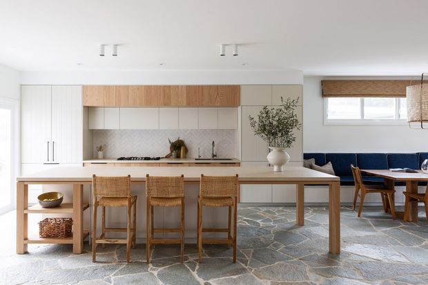 Bright open-plan kitchen and dining area with white cabinetry, warm timber overhead joinery, a long island with integrated seating, woven rattan stools, and a timber dining table. The room features a patterned stone-look floor, white walls, natural textures, and a large window with a woven blind.