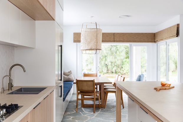 Bright open-plan kitchen and dining area with light timber cabinetry, pale stone benchtops, a large central island, timber dining furniture, woven pendant lighting, and expansive windows overlooking greenery.