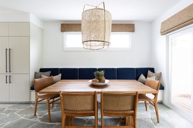 Bright dining area with a long natural timber dining table, upholstered timber dining chairs, and a built-in navy banquette. White walls and cabinetry, woven Roman blinds, a large woven pendant light, and a patterned grey rug create a warm, coastal-inspired setting. Glazing and a light-filled outdoor view are visible on the right.
