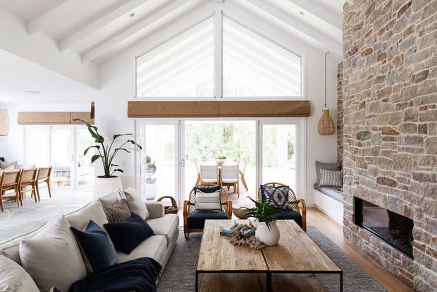 Bright, airy open-plan living room with a large upholstered sectional sofa, timber coffee table, woven rug, and dark accent cushions. The room features a tall pitched ceiling with exposed painted beams, expansive glazed doors and windows, a prominent natural stone fireplace wall, and a dining area visible beyond.