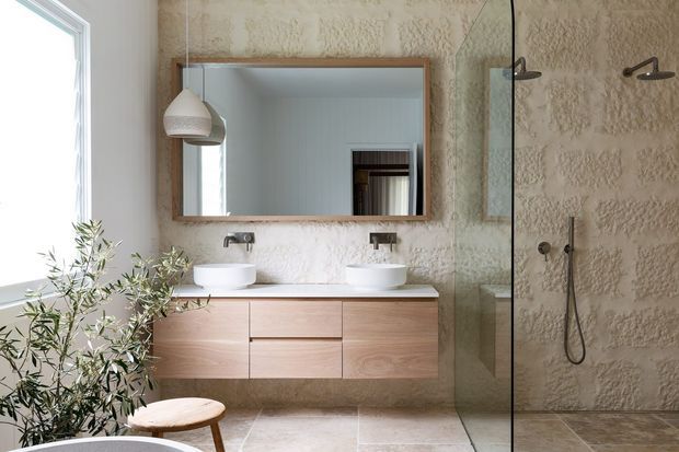 Contemporary bathroom featuring a floating light-wood vanity with two white vessel basins, a large timber-framed mirror, and a frameless glass walk-in shower. The walls have a textured natural stone or plaster finish, with pale stone-look floor tiles, black wall-mounted fixtures, and abundant natural light from a side window. A leafy plant and small timber stool add organic warmth.