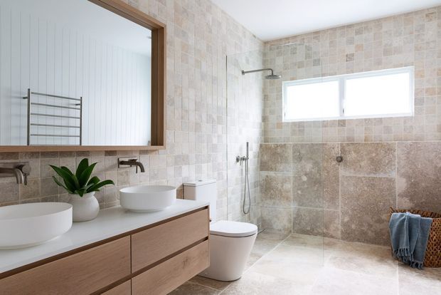 Bright contemporary bathroom with a floating timber vanity, two white vessel basins, wall-mounted brushed metal taps, a large framed mirror, and a walk-in tiled shower with a wall-mounted showerhead. The walls and floor are finished in large-format warm beige and grey stone-look tiles, with a horizontal frosted window providing natural light. A towel ladder, plant, and woven basket add practical and organic touches.