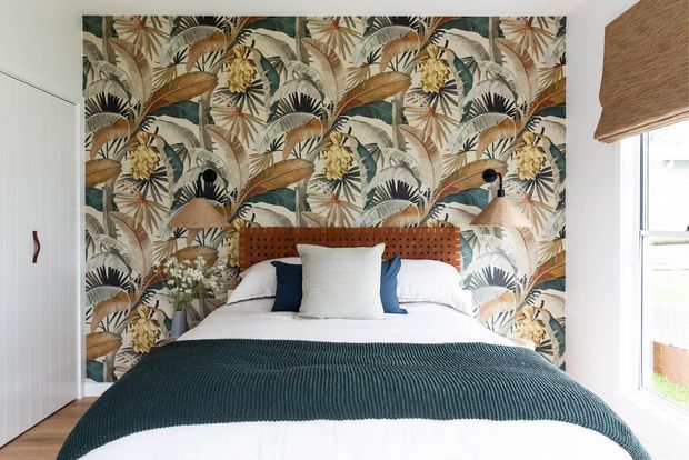A compact, warmly styled bedroom centered on a neatly made bed with a deep teal textured coverlet, white bedding, and layered white, navy, and pale blue pillows. A large tropical botanical wallpaper feature wall spans behind the bed, complemented by a rust-toned woven or upholstered buttoned headboard and two wall-mounted brass-toned reading lights. White vertical-panel built-in cabinetry is visible to the left, while a window with a woven roller blind is on the right. The room has a bright, refined, nature-inspired appearance.