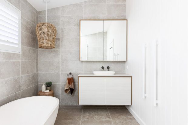Bright contemporary bathroom featuring a freestanding white bathtub, wall-mounted double vanity with integrated basin, mirrored cabinet, large-format grey stone-look wall and floor tiles, white vertical wall panelling, and a woven pendant light. A window with white shutters provides natural light.