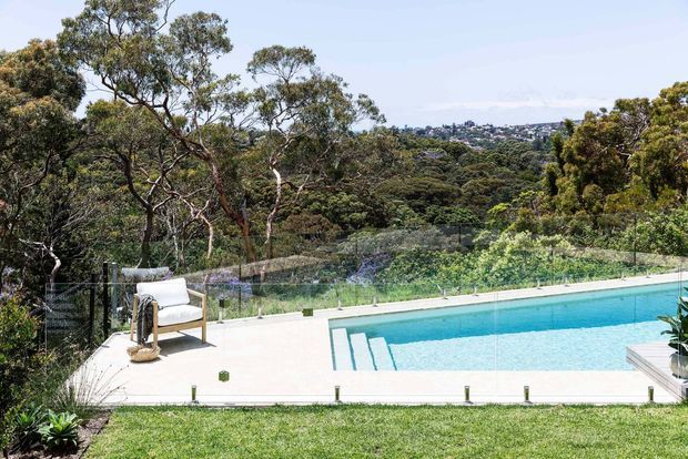 A bright outdoor swimming pool terrace set within a landscaped garden, with a clear glass safety balustrade, pale paved pool surround, submerged entry steps, and a single outdoor lounge chair. Mature trees and a leafy suburban hillside are visible beyond the pool.