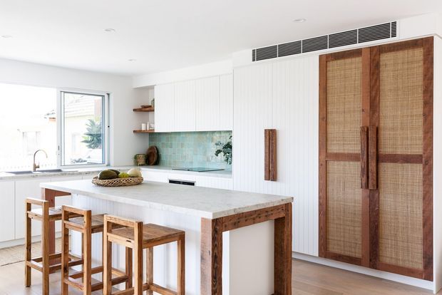 Bright open-plan kitchen featuring a large white island with timber detailing and four timber stools, white vertical wall panelling, a pale aqua patterned tile splashback, white cabinetry, and a tall woven rattan timber-framed storage door. Natural light enters through a large window, with warm timber flooring and accents creating a relaxed coastal feel.