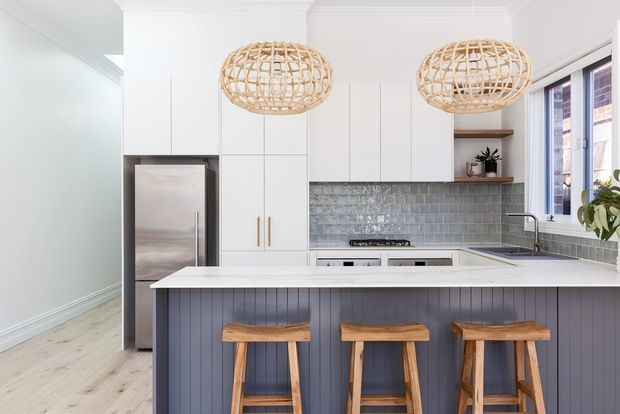 Bright contemporary kitchen with white full-height cabinetry, a stainless-steel refrigerator, grey subway-tile splashback, white benchtops, and a dark blue-grey vertical-panelled island with three timber stools. Two woven rattan pendant lights hang above the island, with timber flooring and a large window adding natural light.