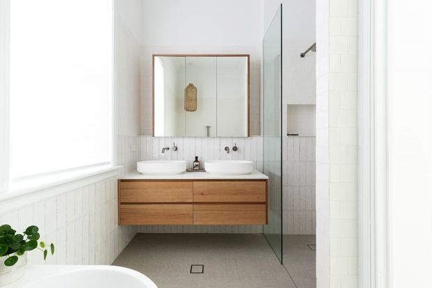 Bright contemporary bathroom with a floating mid-tone timber vanity, twin white vessel basins, wall-mounted brushed metal tapware, a large framed mirror, white vertical wall tiling, and a frameless glass shower enclosure. A freestanding bathtub is partially visible in the foreground, with a small plant adding greenery.