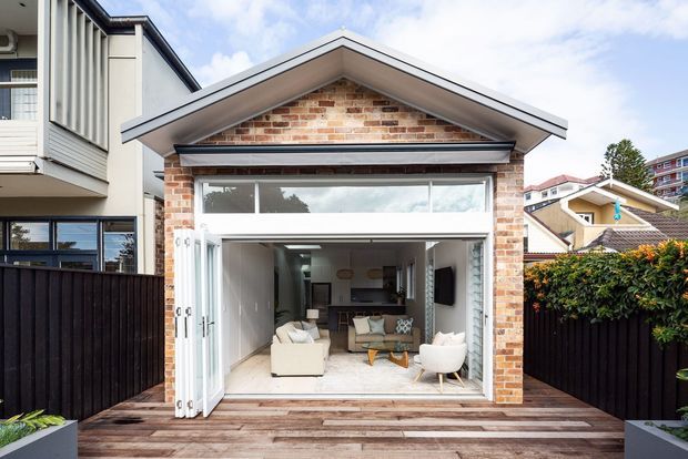 A compact contemporary rear extension or pavilion with a prominent front-facing gable roof, exposed red brick facade, wide clerestory glazing, and fully opened bi-fold doors. A timber deck leads into a bright living room with seating and a visible kitchen area. The building is tightly framed by neighbouring residences and dark fencing.