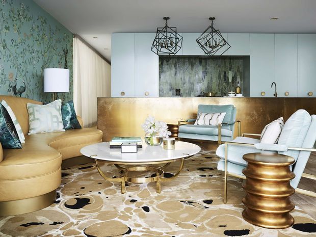 A glamorous open-plan living room with a curved tan sectional sofa, pale blue upholstered lounge chairs, a round white coffee table, and a large abstract patterned rug. The room includes a compact kitchen at the rear with pale blue slab cabinetry, a metallic bronze-gold island or backsplash surface, and a dark textured feature wall. Botanical wallpaper, sheer curtains, geometric pendant lights, brass-toned accents, and sculptural furniture create a highly decorative, luxurious interior.
