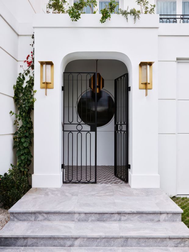 Front exterior entry of a refined white-rendered residence, featuring a deep arched porch, decorative black metal security gates, brass wall sconces, pale stone steps and paving, and climbing greenery. A small upper-level balcony with planting and black railing is visible above.
