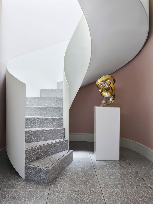 A sculptural curved stairway with a sweeping white balustrade and grey terrazzo steps, set against a muted dusty-rose wall. Large grey floor tiles continue through the space, while a gold abstract sculpture sits on a white plinth beside the stairs. The composition is refined and gallery-like, with strong flowing architectural curves and a restrained palette.