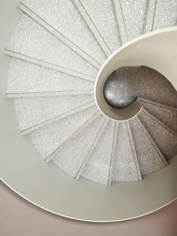 Top-down view of a sculptural spiral staircase curving around a central circular void. The stair treads have a speckled light finish with fine warm-toned edging, while the inner curved balustrade or wall is smooth warm grey-beige. A darker speckled stair surface is visible through the opening, creating a striking geometric spiral composition.