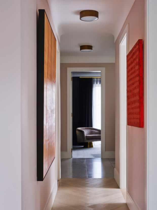 A narrow, well-finished residential hallway viewed toward a living room. The corridor has pale pink-beige walls, light herringbone timber flooring, white door and architrave trim, and two flush-mount cylindrical ceiling lights with dark wood-toned surrounds. A large vertical timber-framed artwork or panel lines the left wall, while a vivid red textured artwork hangs on the right. At the far end, a dark-curtained room with a leather armchair and patterned rug is visible.