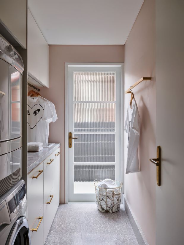 A narrow, compact laundry room with white built-in cabinetry and brass handles, stacked laundry appliances along the left wall, a pale stone-look benchtop, and hanging rails for clothing. A frosted fluted-glass door provides diffused daylight at the end of the room. The space has soft blush-pink walls, a white ceiling, and a light grey textured floor, with a wire laundry basket in the centre.