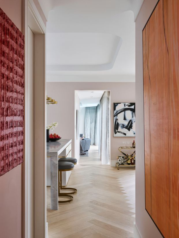 A refined interior view looking through a hallway toward a living room. The space features pale herringbone timber flooring, soft taupe walls, a sculptural recessed ceiling detail, a stone-topped peninsula with dark upholstered stools, a large ribbed timber wall panel, contemporary artwork, and layered views into the living area. The palette is warm and sophisticated with burgundy, timber, cream, and muted neutral tones.