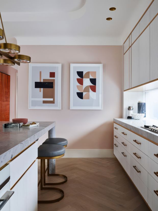 A refined contemporary kitchen with a central island and two curved black stools, pale pink walls, white slab-front cabinetry, warm timber trim, marble-look benchtops, and integrated under-cabinet lighting. The floor is laid in a warm herringbone timber pattern, while geometric framed artwork and sculptural brass pendant lighting add decorative character.
