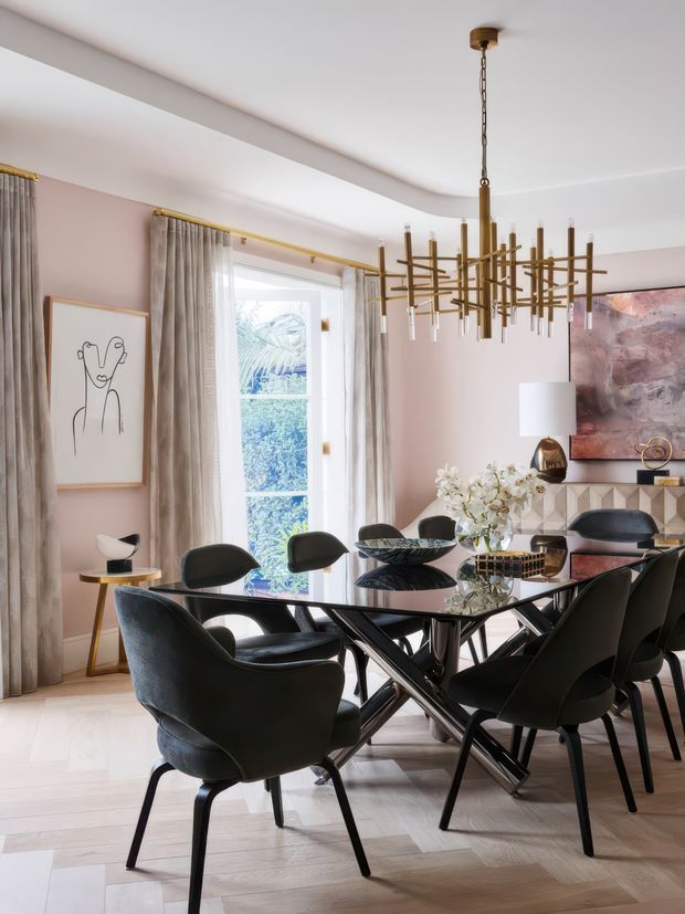 A refined dining room with a glossy black glass dining table surrounded by sculptural black dining chairs. The room features pale pink walls, light timber flooring, full-height drapery, a large geometric brass chandelier, contemporary artwork, and a decorative sideboard with geometric patterned doors. Natural light enters through tall windows, creating an elegant, high-contrast interior.