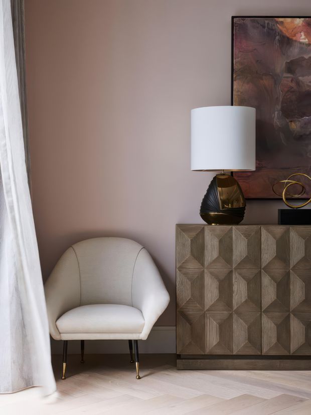 A refined interior vignette featuring a sculptural cream upholstered armchair beside a geometric dark wood sideboard. A sculptural table lamp, abstract artwork, and brass decorative object sit against a warm taupe wall, with sheer curtains and pale herringbone timber flooring adding softness and texture.