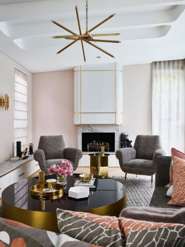 Elegant contemporary living room with a sculptural white ceiling, curved fireplace surround, pale blush walls, and a large brass starburst pendant. The room features two textured grey lounge chairs, a curved sofa with patterned cushions, a glossy black and brass round coffee table, white built-in cabinetry, sheer curtains, and a marble-look fireplace surround.