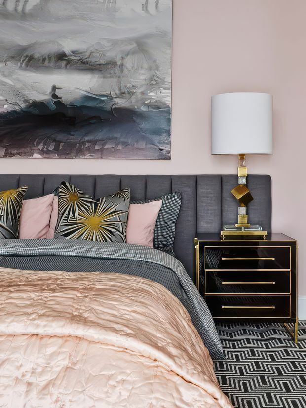 A stylish bedroom featuring a tall charcoal upholstered bedhead, layered blush pink and patterned bedding, and a black-and-gold geometric bedside table with a sculptural lamp. A large abstract artwork hangs above the bed, while a bold black-and-white geometric rug adds contrast. The room has a refined, glamorous contemporary aesthetic with soft pink, charcoal, black, white, and gold accents.